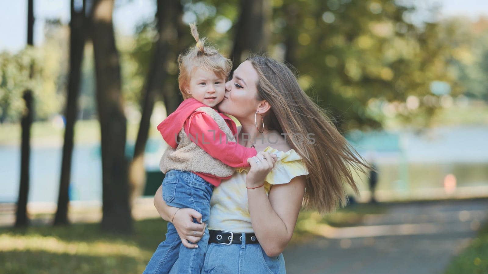 Young mother kissing her daughter on the cheek while joyfully holding her in her arms, surrounded by the beauty of a sunny park