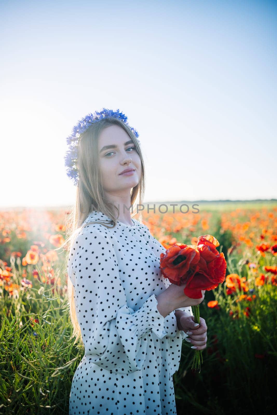 Blonde woman wearing floral headpiece, holding poppy bouquet while standing in sunlit meadow, radiating natural beauty and summer warmth