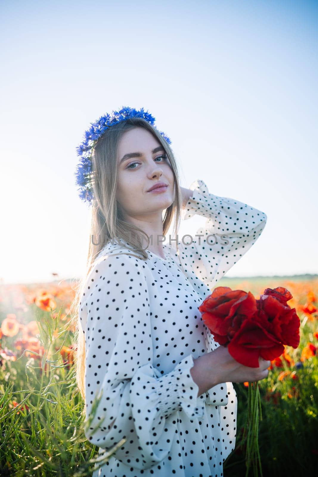 Beautiful blonde woman with cornflower wreath holding a bouquet of poppies in a field during a sunny summer day