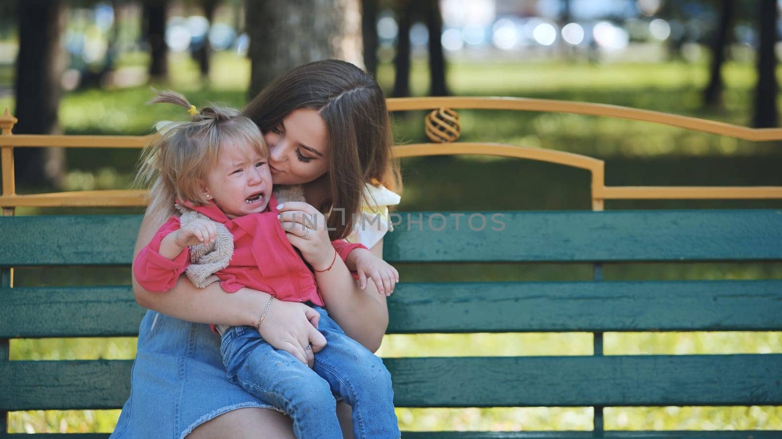Mother soothing tearful toddler while sitting on park bench under bright sunlight, offering comfort and reassurance