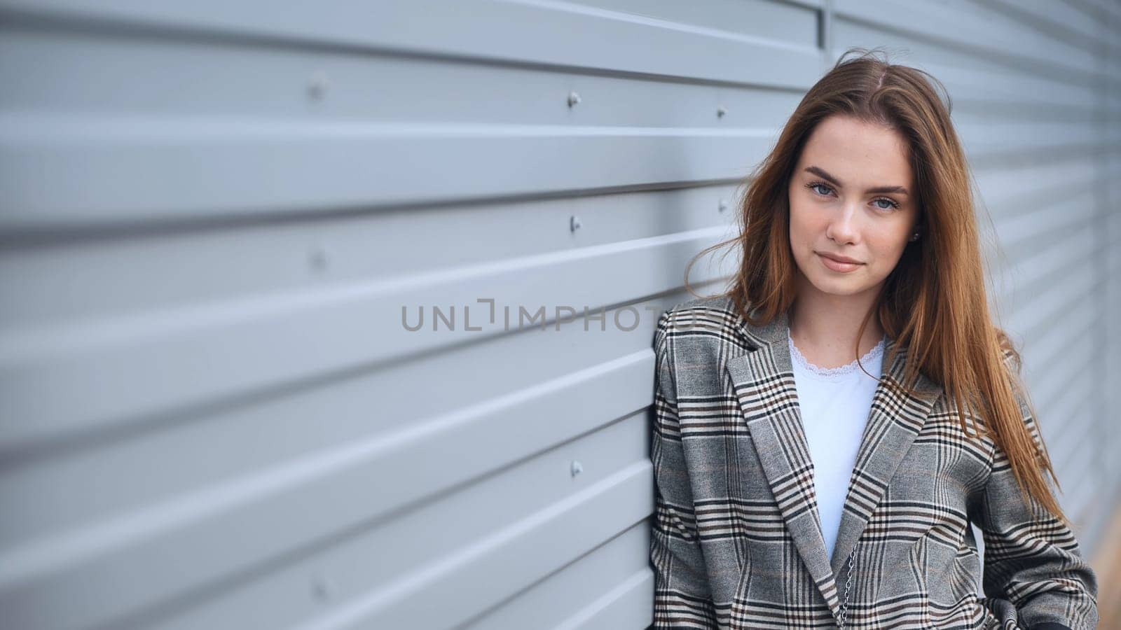 Confident businesswoman leaning against gray metal wall, displaying professional urban style with strong posture and self assured expression