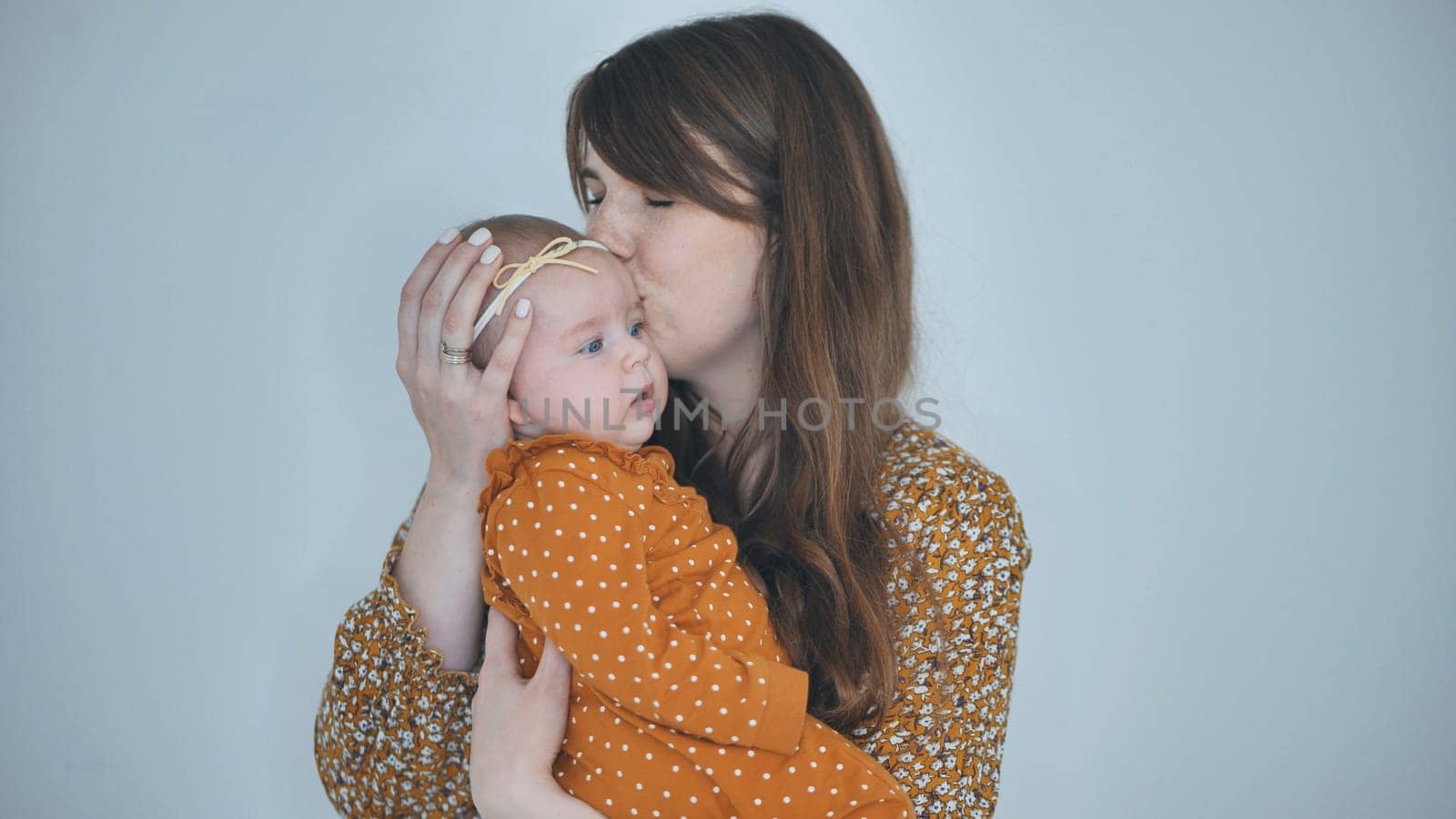 Studio portrait of a happy mother kissing her baby girl on the forehead, holding her in arms, both wearing orange clothes