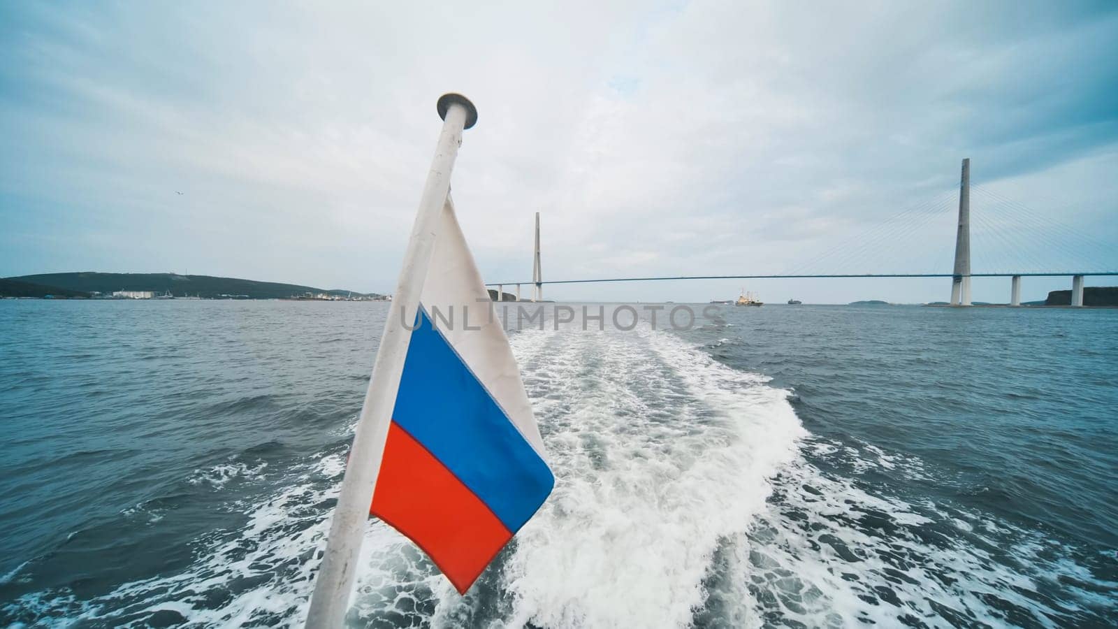 Sailing towards russky bridge, a boat glides through the ocean, with the russian flag proudly waving in the summer breeze under a cloudy sky