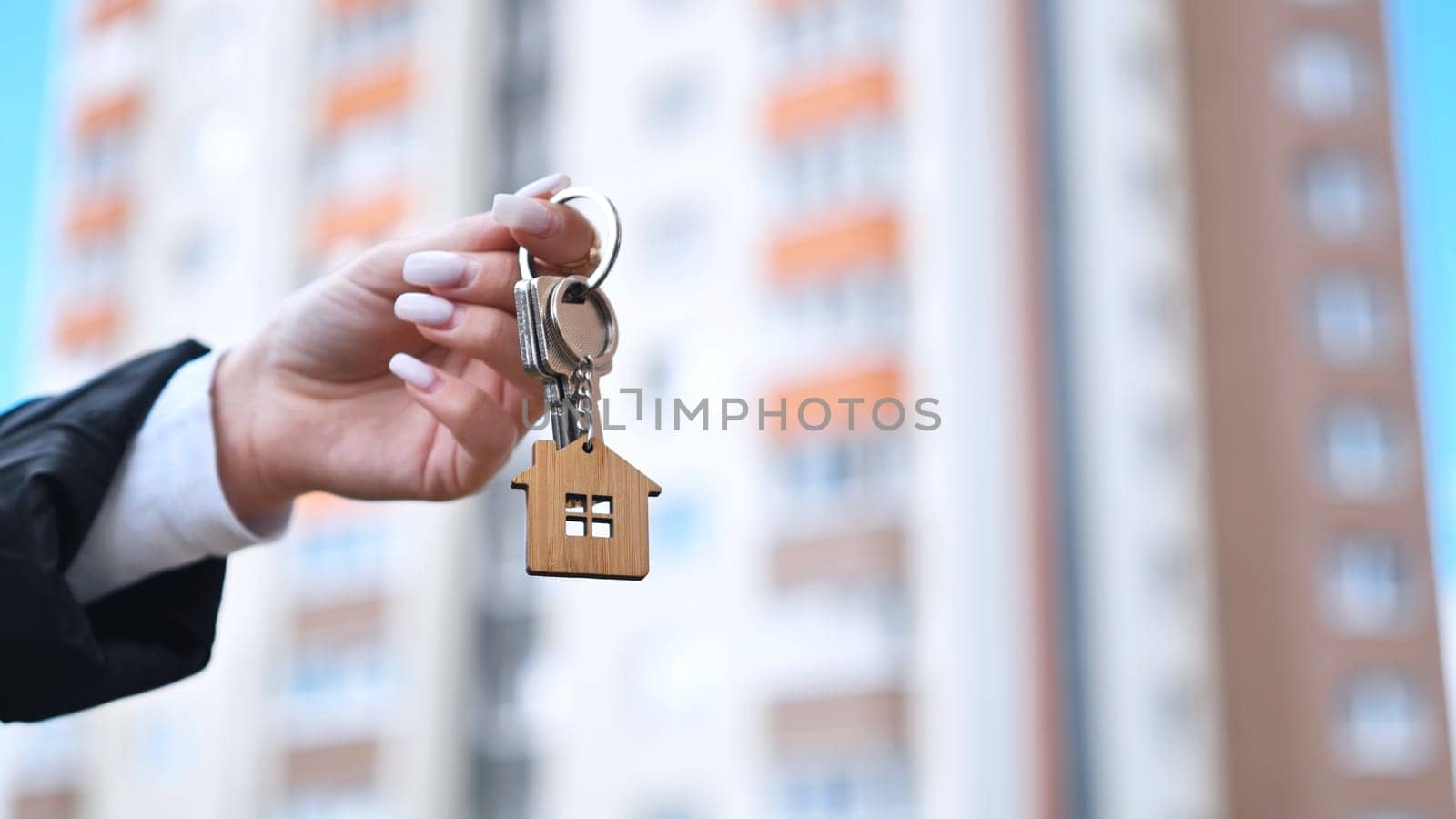 Realtor holding house shaped wooden keychain, standing near modern residential complex, representing property sale