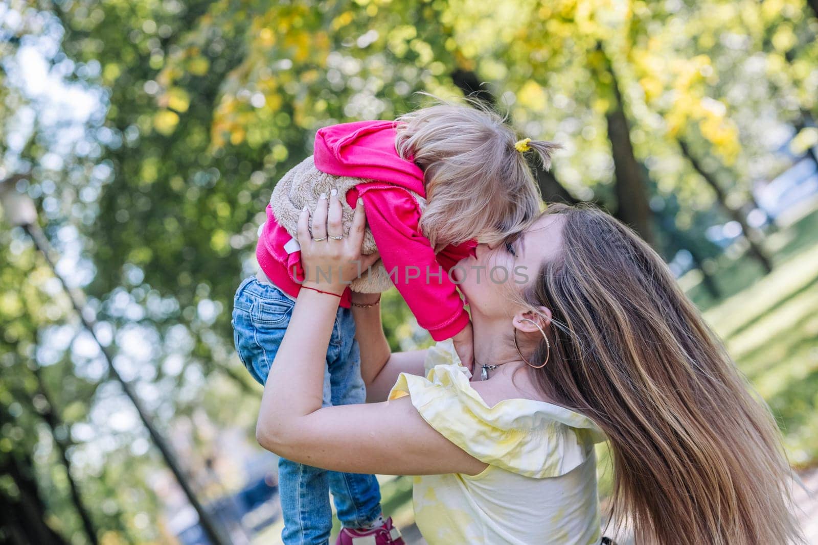 Mother holding her daughter in her arms, sharing a tender kiss while enjoying a sunny day together in a vibrant park filled with greenery