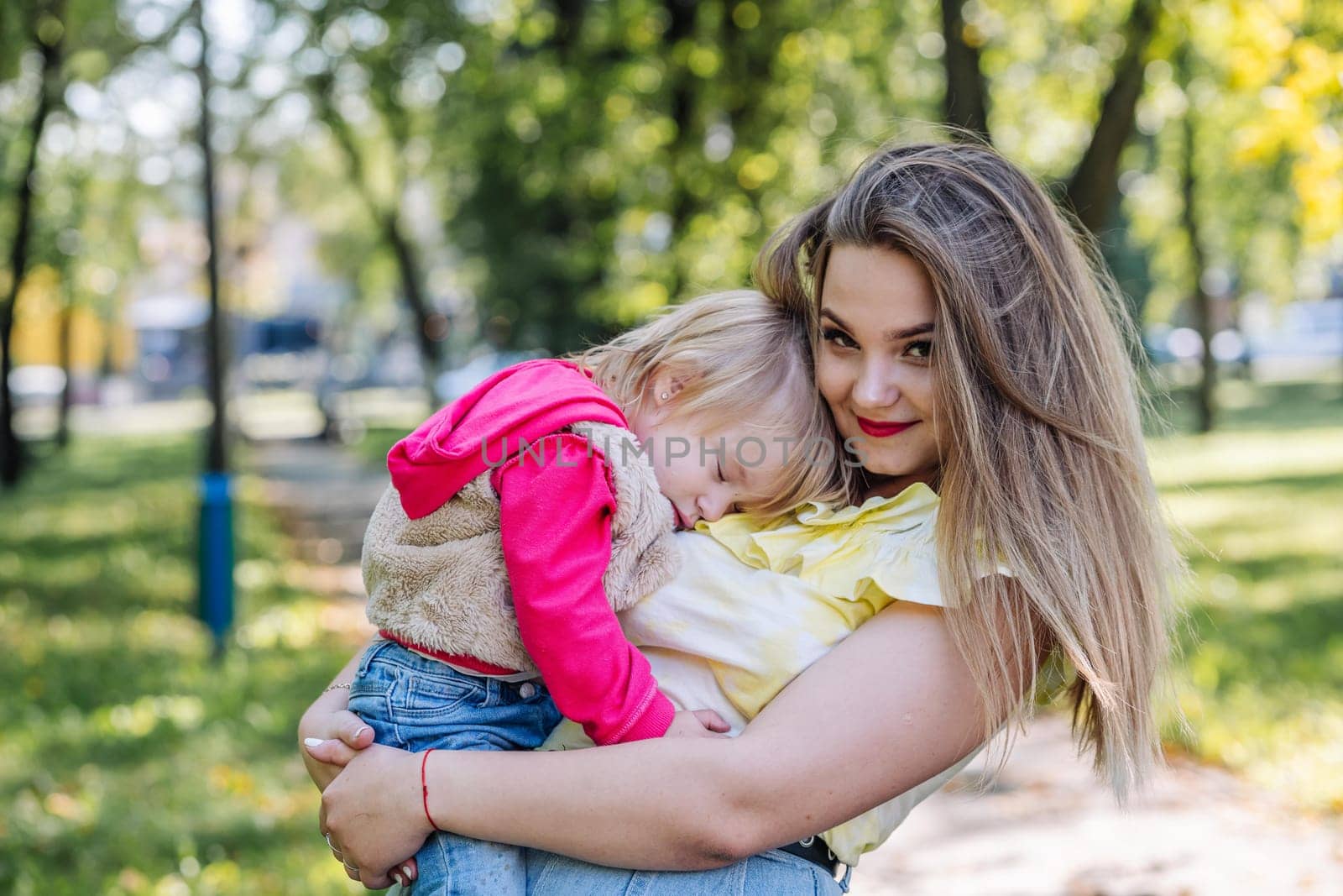 Mother cuddling peacefully sleeping daughter, sunlight filtering through verdant park landscape, tender maternal moment unfolding