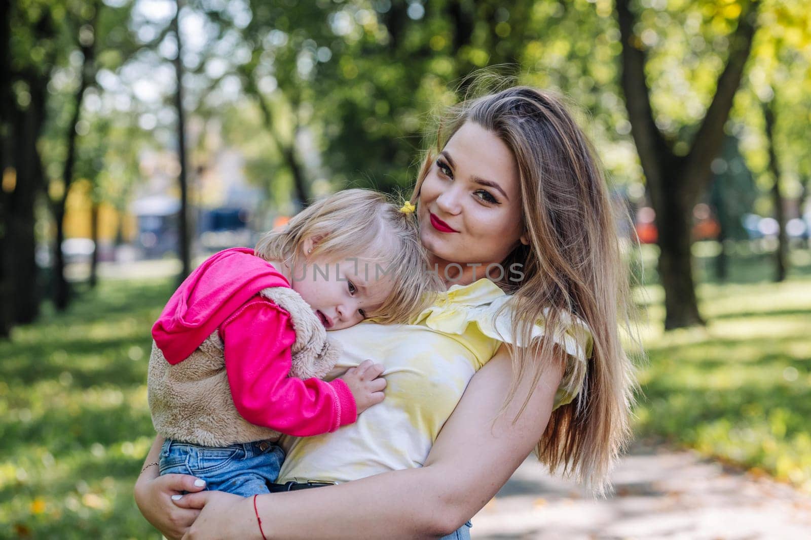 Young mother holding her sad little daughter in her arms, providing comfort and love in a sunny park filled with greenery and warmth