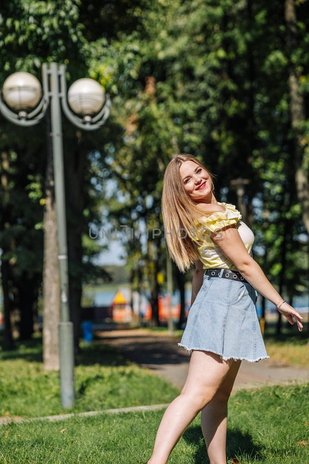 Blonde woman wearing a yellow top and denim skirt smiling and walking in a park during a sunny day