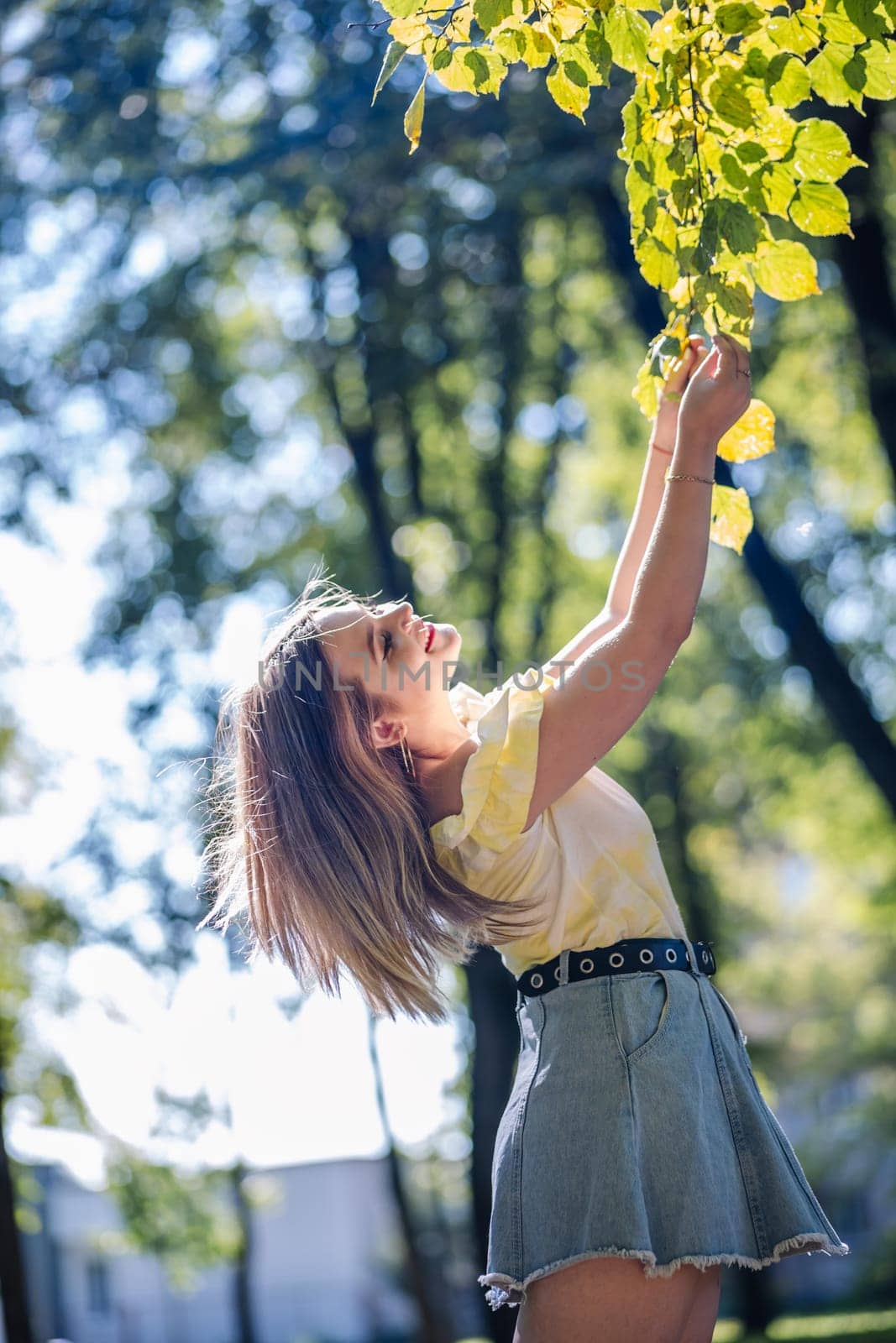 Teenager enjoying a sunny day in a park, reaching for leaves on a tree branch, feeling the warmth of nature