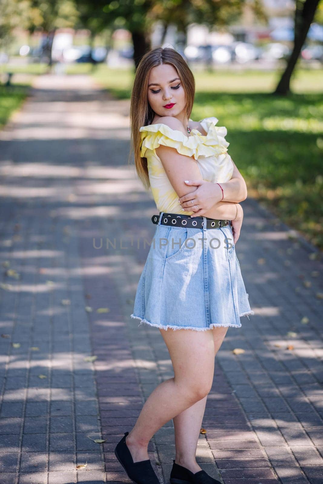 Fashionable young woman wearing a ruffled yellow top and denim skirt posing in a park during a sunny summer day