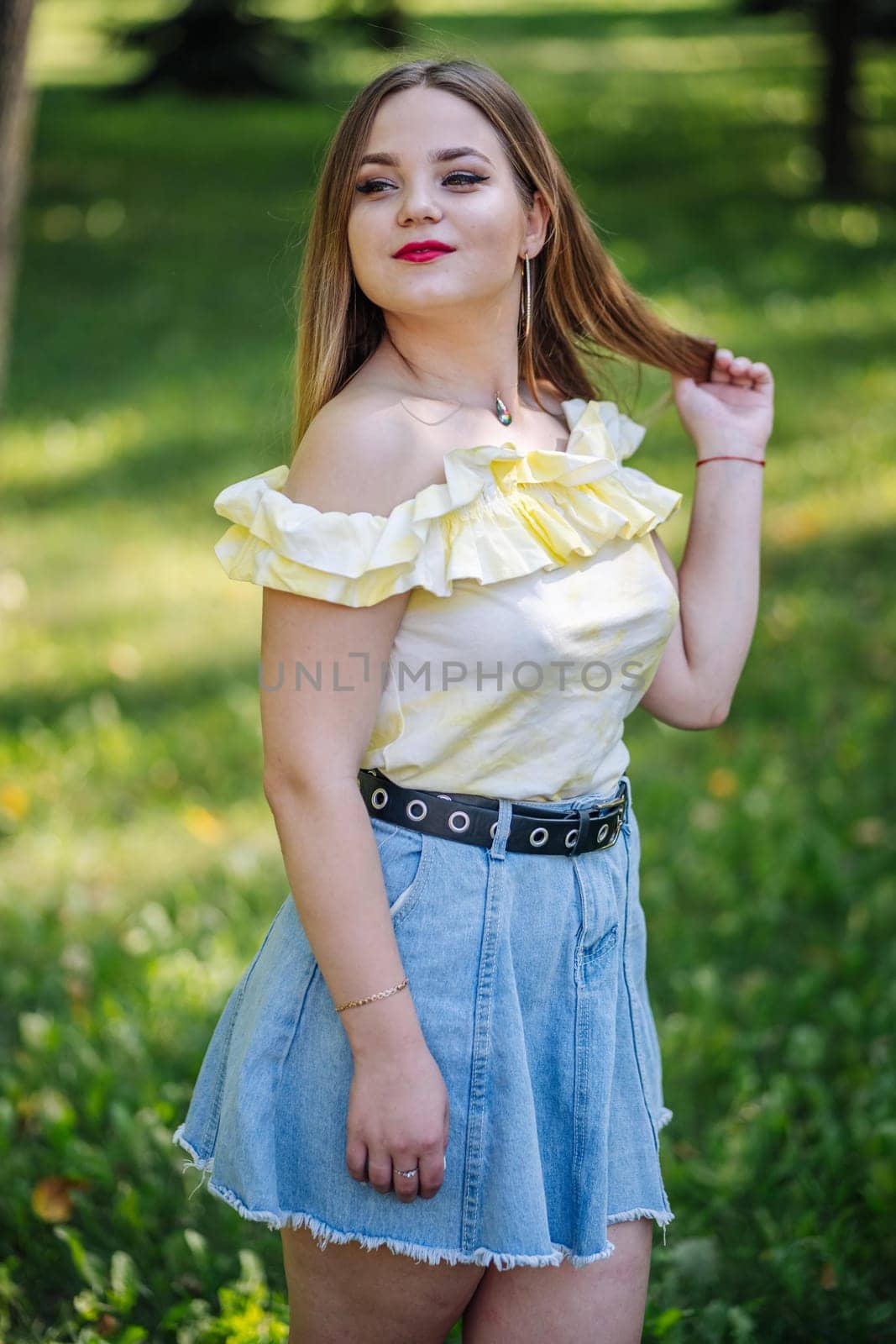 Stylish female with blonde locks, sporting yellow blouse and denim skirt, radiating confidence while standing in lush green park setting