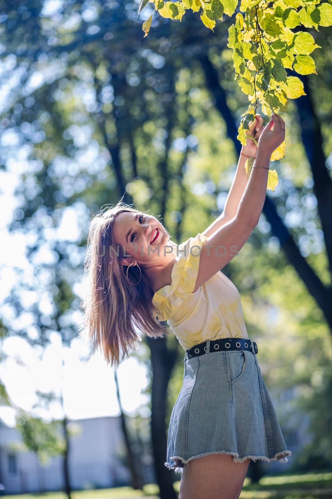 Teenager girl with long hair, wearing a yellow t shirt and a jeans skirt, is enjoying a sunny day in a park, touching the leaves of a branch