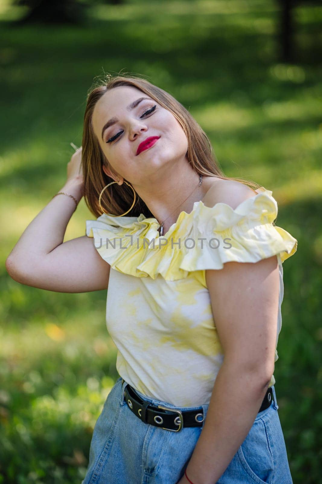 Fashionable young woman adjusting her hair while posing in a park, wearing a stylish summer outfit