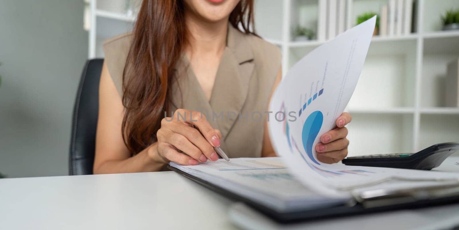 An office worker meticulously analyzes performance reports, holding a pen for notes, displaying a focus on business metrics and analysis in a modern workspace.