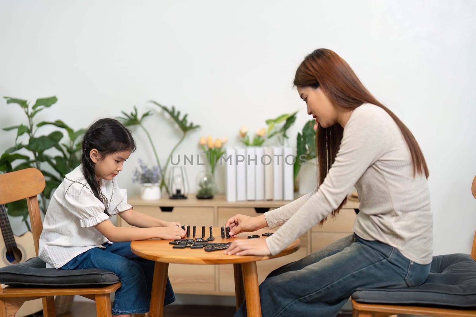 The scene depicts a calm moment where the mother and daughter are engaged in a strategic game of dominos, enhancing their bond through shared experiences.