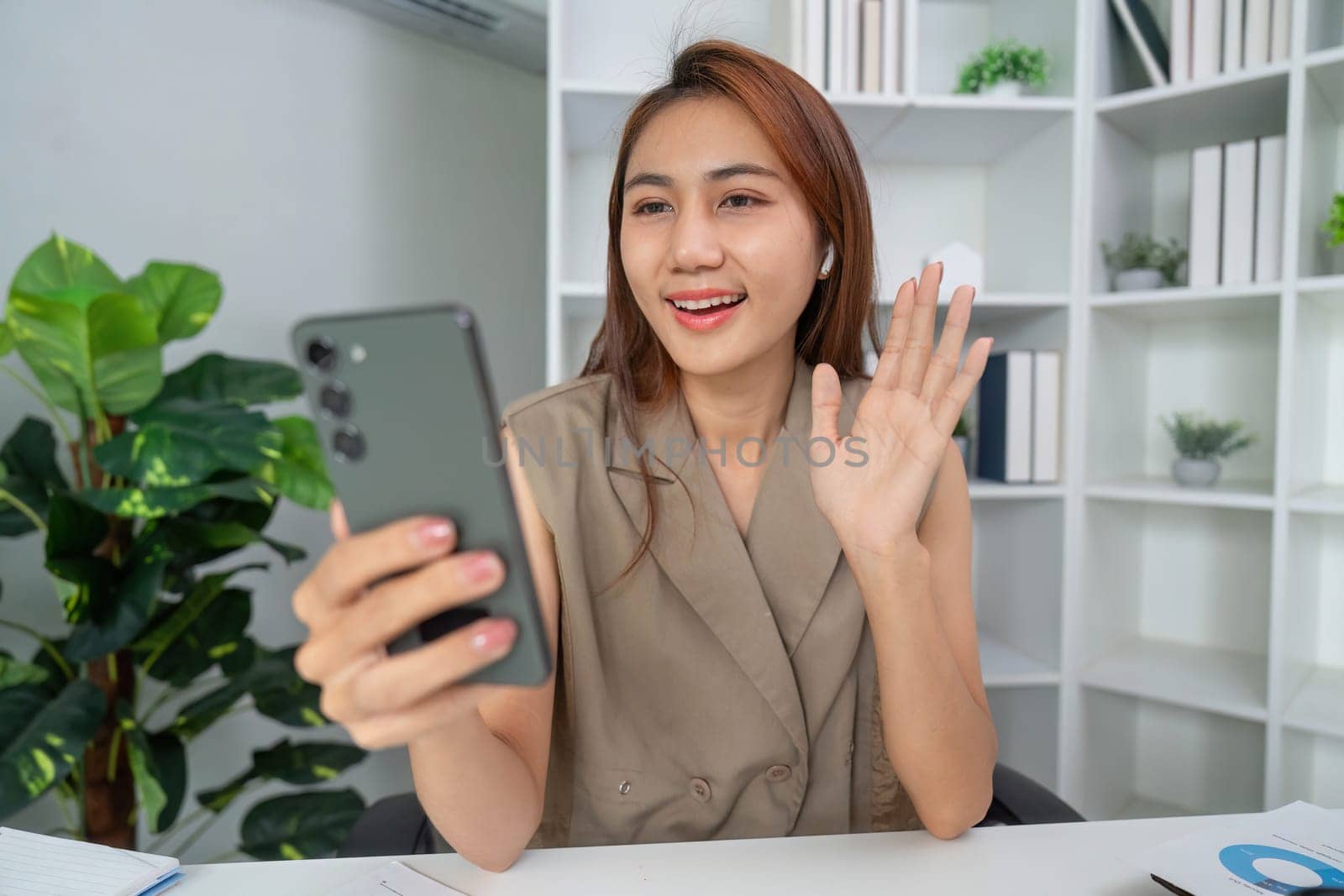A young woman smiles and waves on a video call, showcasing a lively interaction. Her earbuds highlight her engagement in modern communication.
