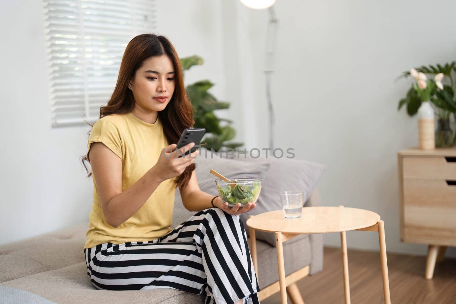 A woman sits on a sofa, checking her phone while having a nutritious salad, showcasing a balance of technology and wellness.