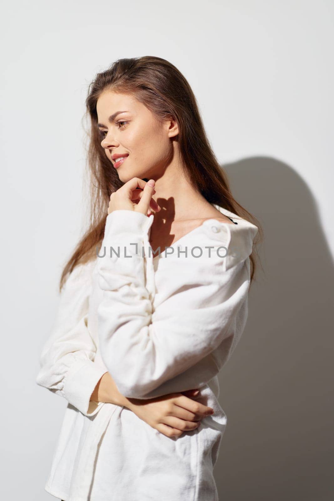 This image features a young woman with long hair, dressed in a chic white blouse, posing thoughtfully against a soft light background The warm lighting accentuates her natural beauty, showcasing a s