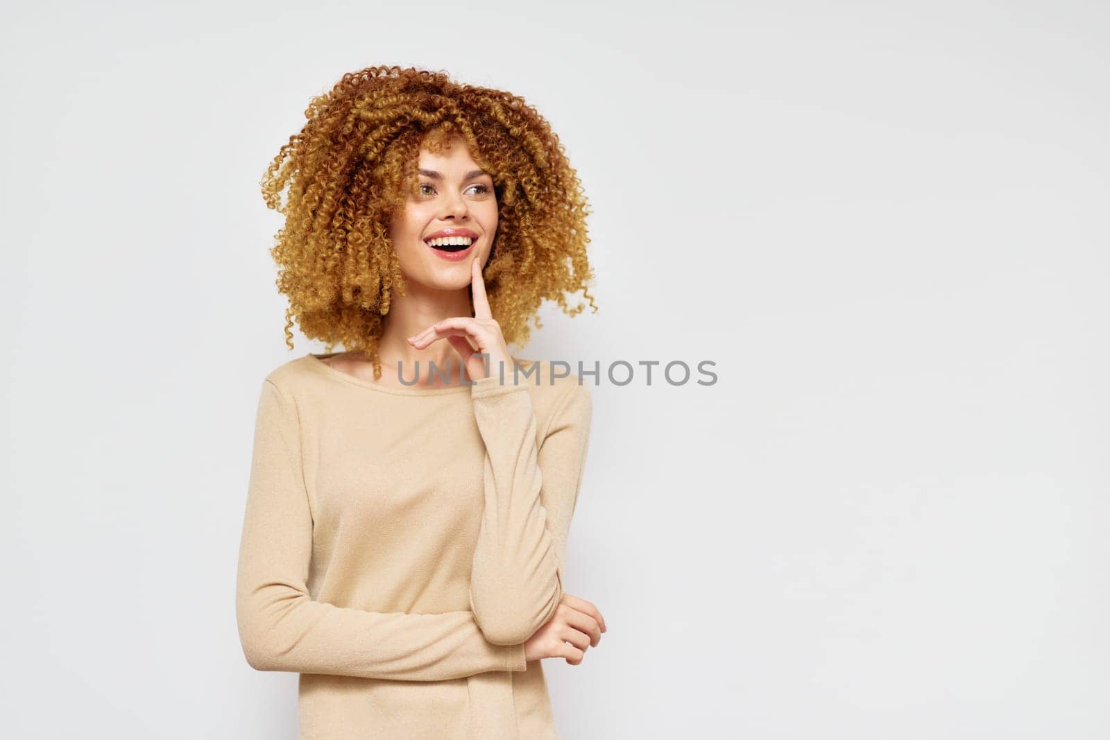 Smiling woman with voluminous curly hair stands against a light gray background, dressed in a beige long sleeve top Her joyful expression conveys happiness and self assuredness, ideal for lifestyle,