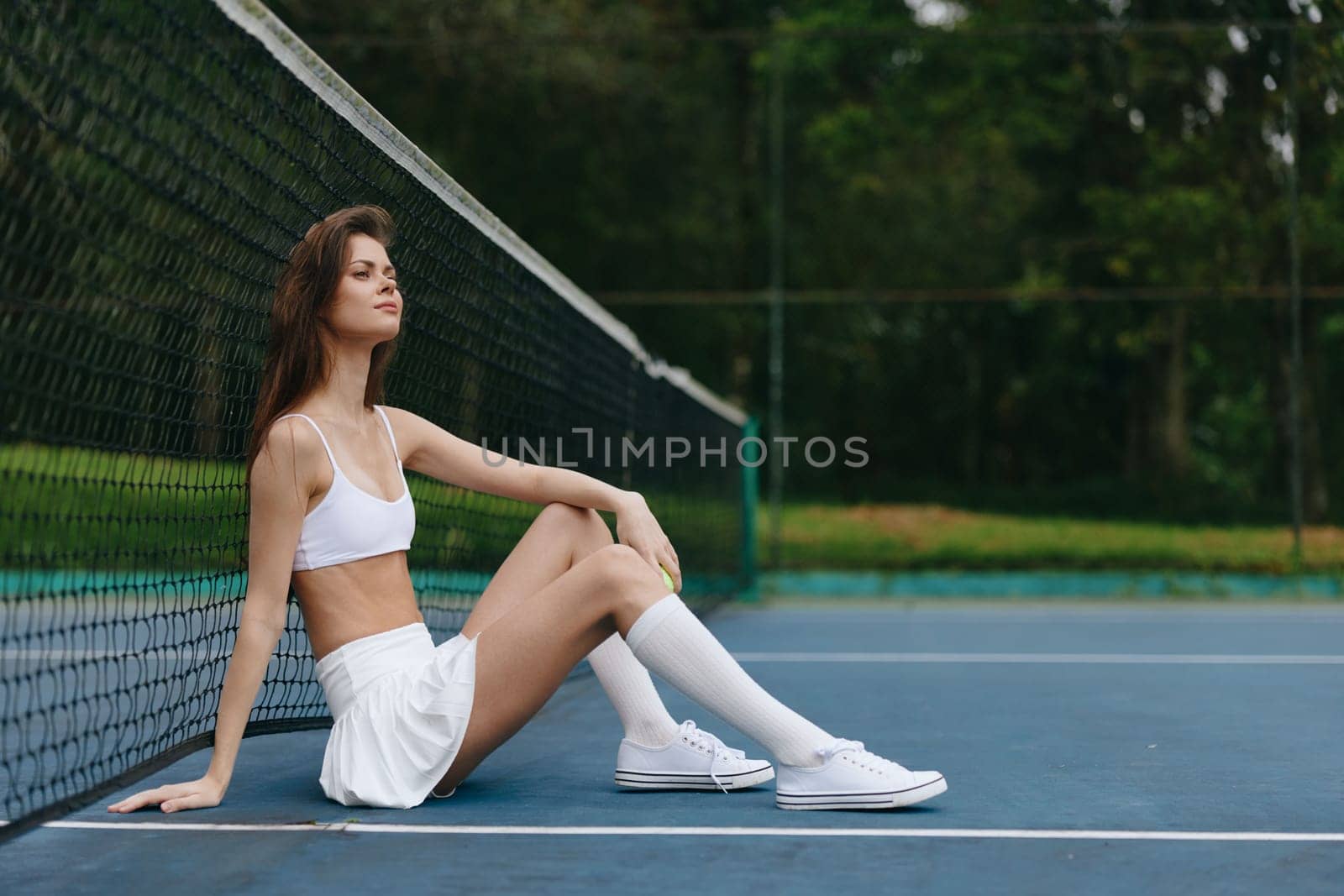 A confident young woman in sportswear relaxing on a tennis court, showcasing joy and a healthy lifestyle through her athletic pose and serene environment