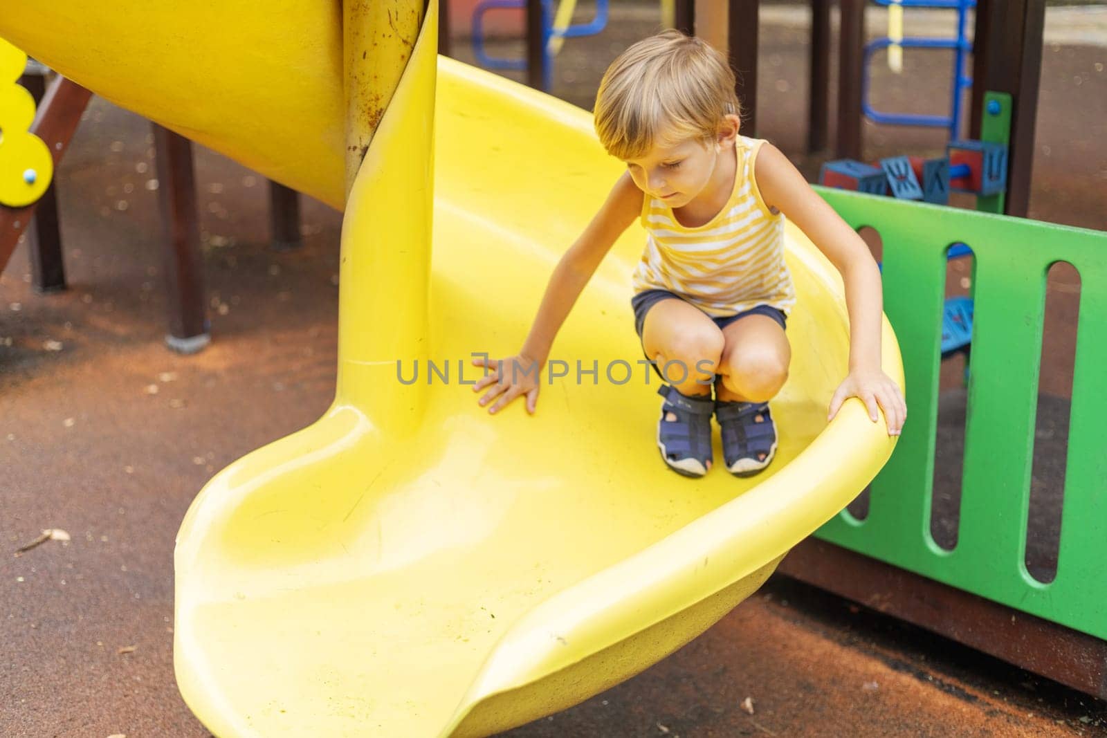Boy wearing striped shirt crouching on a yellow spiral slide at an outdoor playground with rubber flooring and colorful barriers