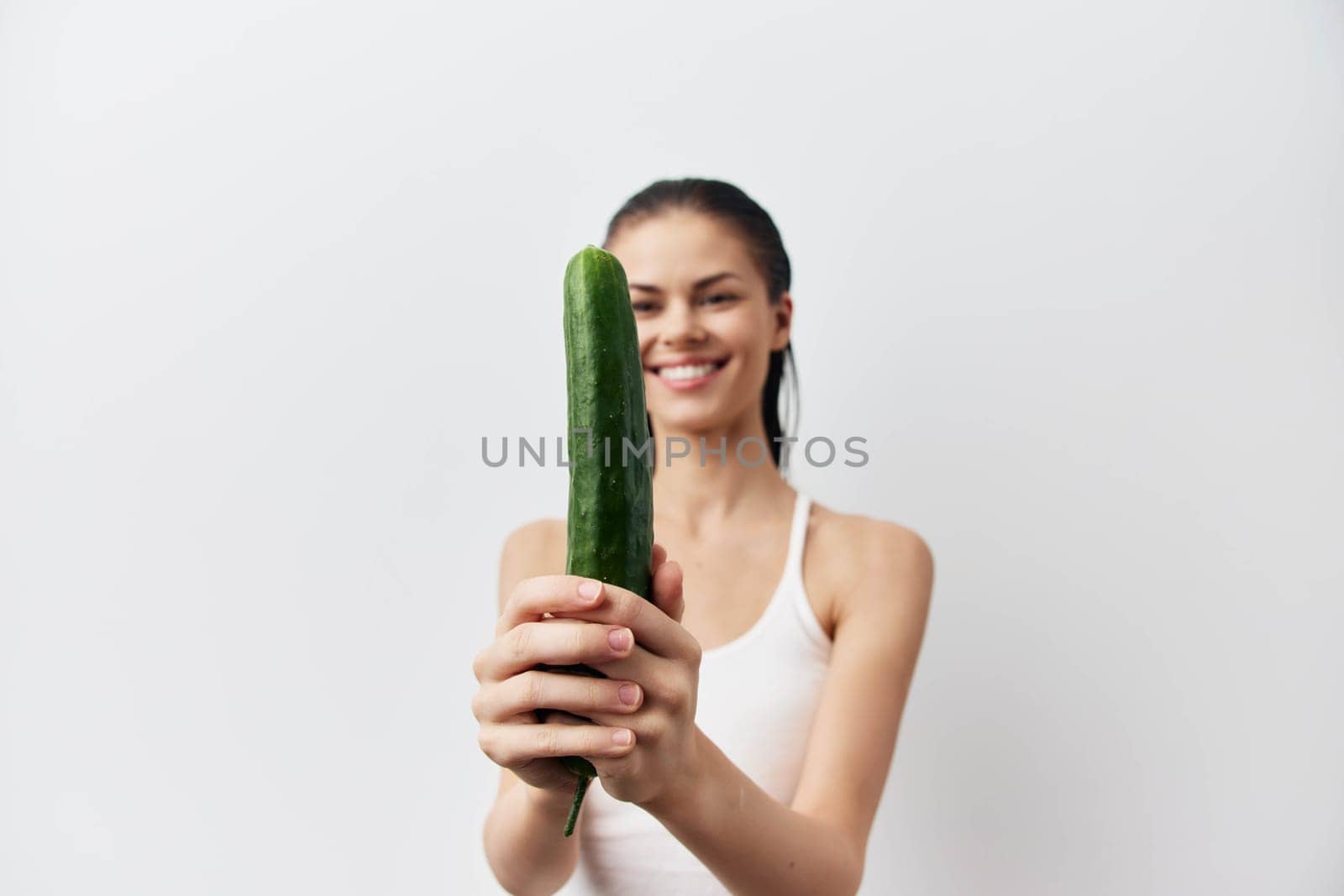 Smiling woman confidently holds a fresh cucumber against a plain white background The image conveys a lighthearted and vibrant atmosphere, promoting healthy eating and wellness