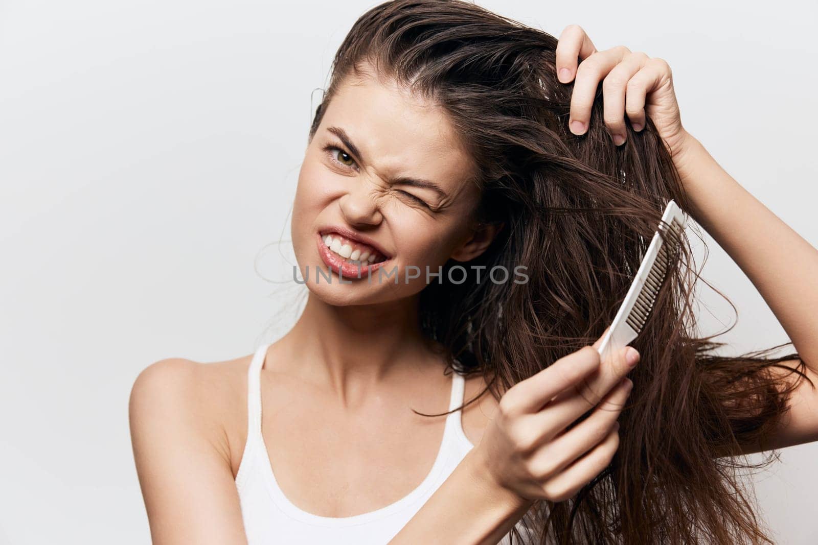 A young woman is shown staring at the camera while using a comb on her long hair, displaying a mix of frustration and determination against a clean white backdrop, capturing relatable hair care moments