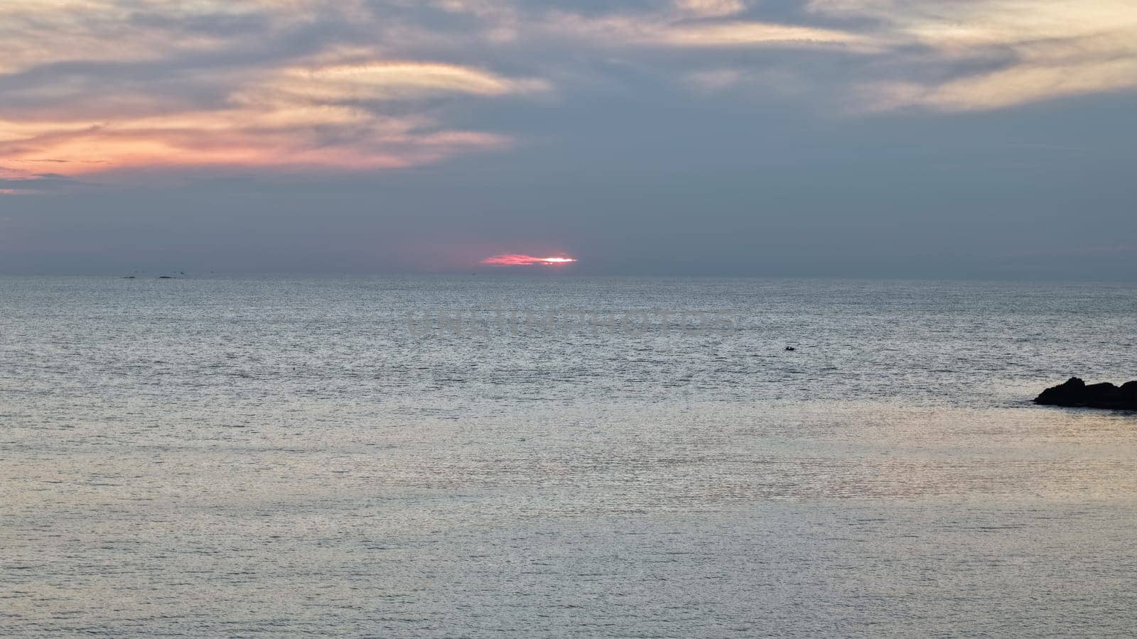 Pastel pink and grey hues blending during sunset, reflecting beautifully on calm ocean waters. Rocks in the foreground enhance the tranquil and serene atmosphere of the scene