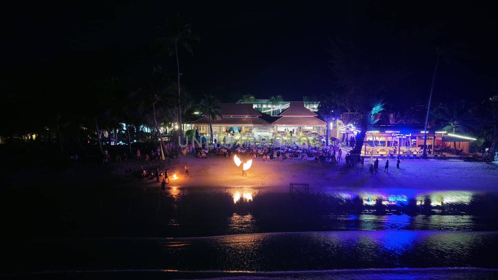 Tourists are relaxing and enjoying a fire show on a beach at night, with restaurants and palm trees in the background, creating a vibrant and tropical atmosphere