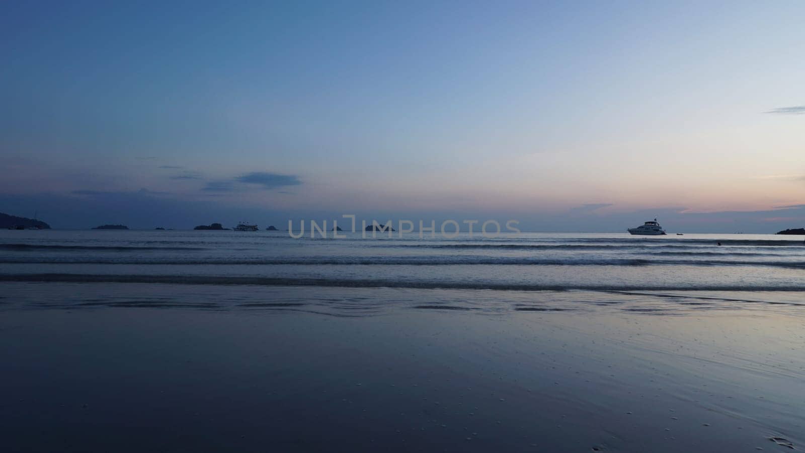 As the sun sets, tourists enjoy the gentle waves of a tropical ocean, with boats dotting the horizon and islands adding a touch of serenity to the tranquil scene