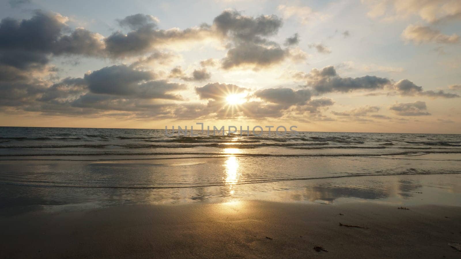 Soft sunlight filters through a cloudy sky, illuminating gentle waves rolling onto a wet sandy beach, creating a serene and reflective seascape at sunset