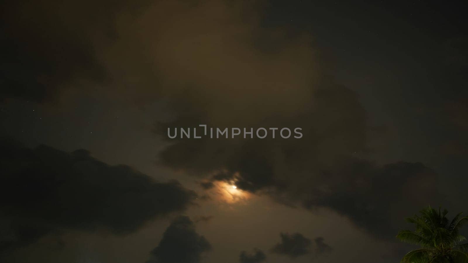 The moon shines through dark clouds at night, creating a moody and atmospheric scene with a few stars visible and a palm tree silhouette in the corner