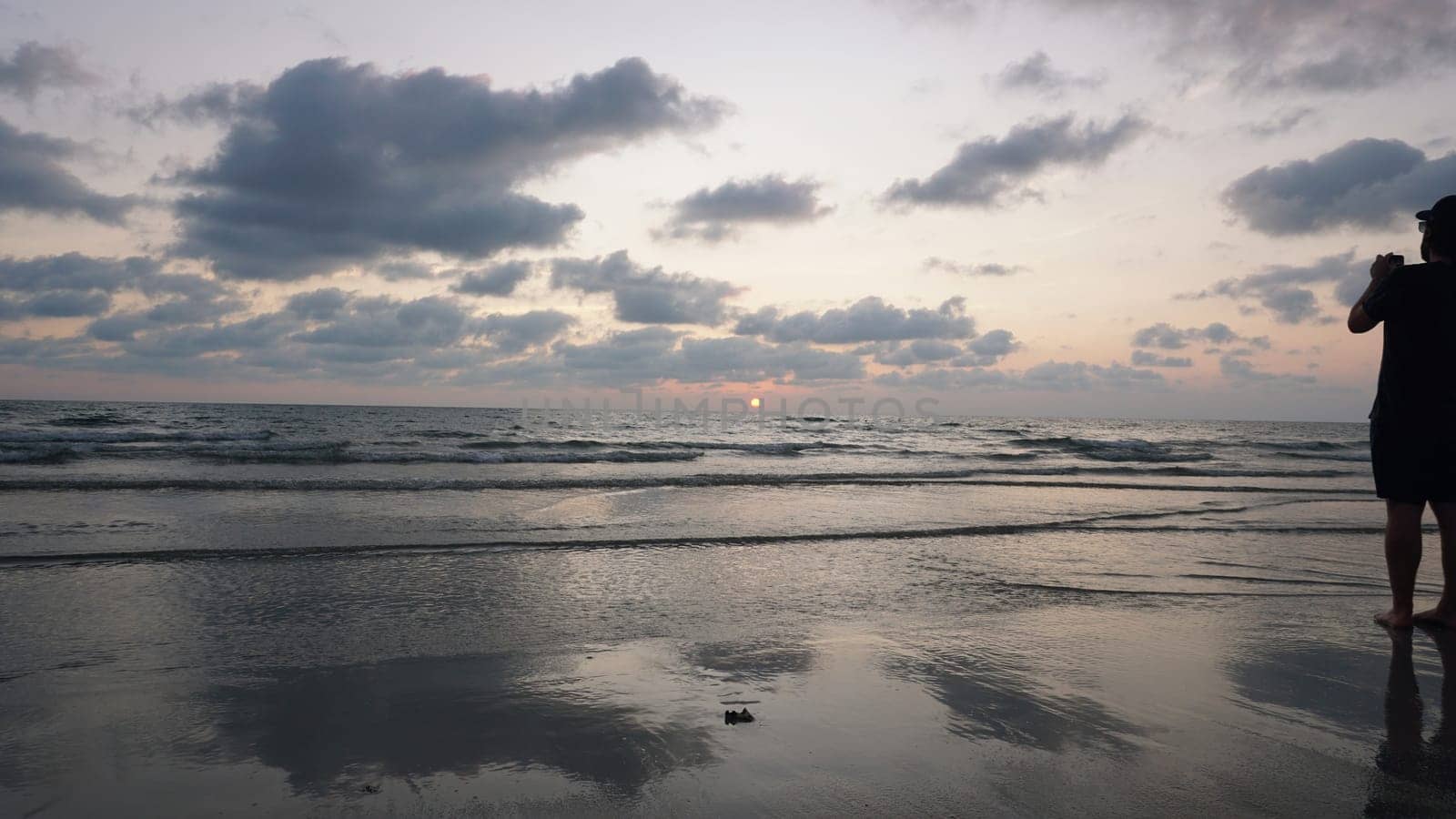 Photographer standing on a wet sandy beach at sunset, capturing vibrant sky colors reflected in gentle waves as the sun sets below the horizon