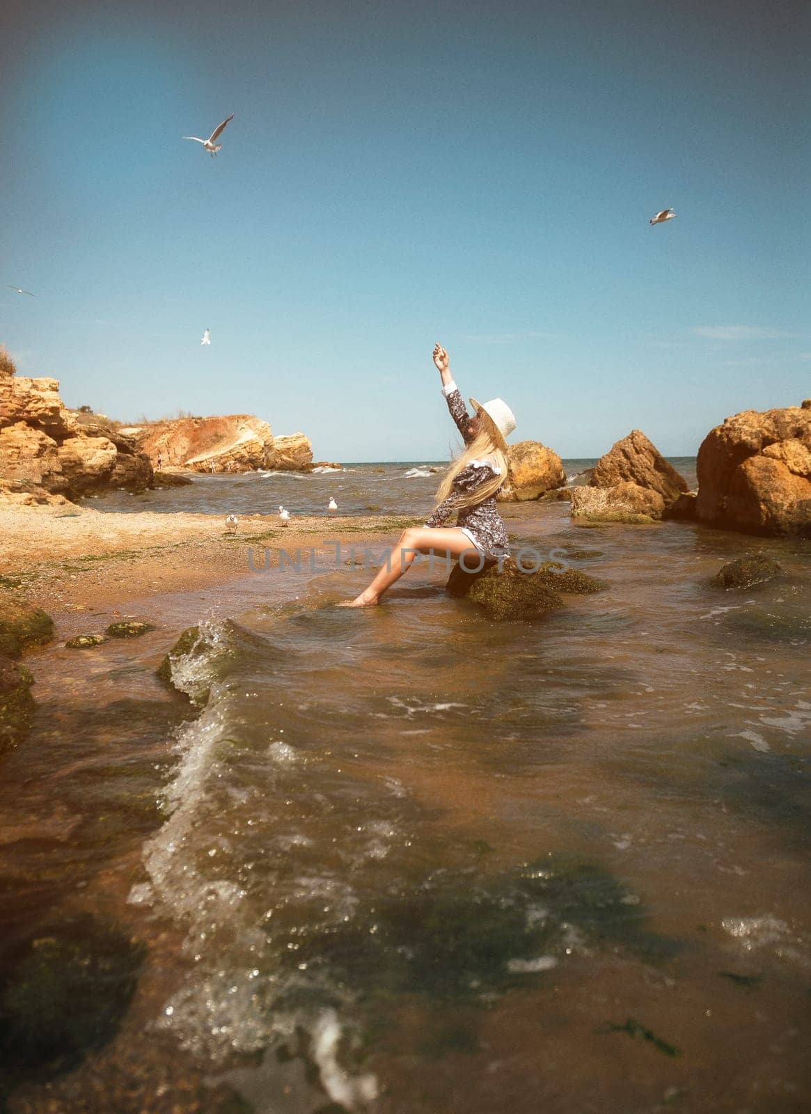young blonde in a short dress and hat sits on a rock in the sea on a deserted beach. her hair flutters in the wind, she enjoys the sun, seagulls fly around her general plan