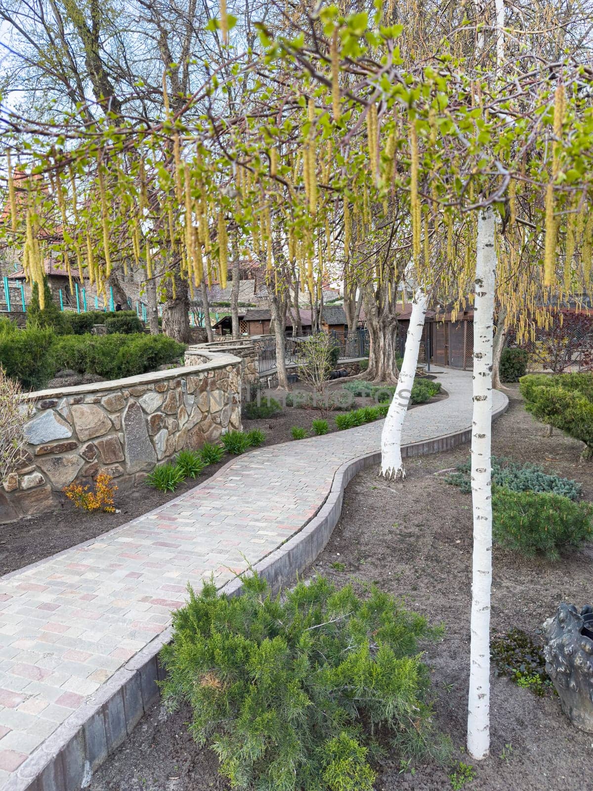 A winding garden path showcases vibrant blooms and lush greenery in a peaceful outdoor area. The landscaping features stone walls and a variety of plants under clear skies.
