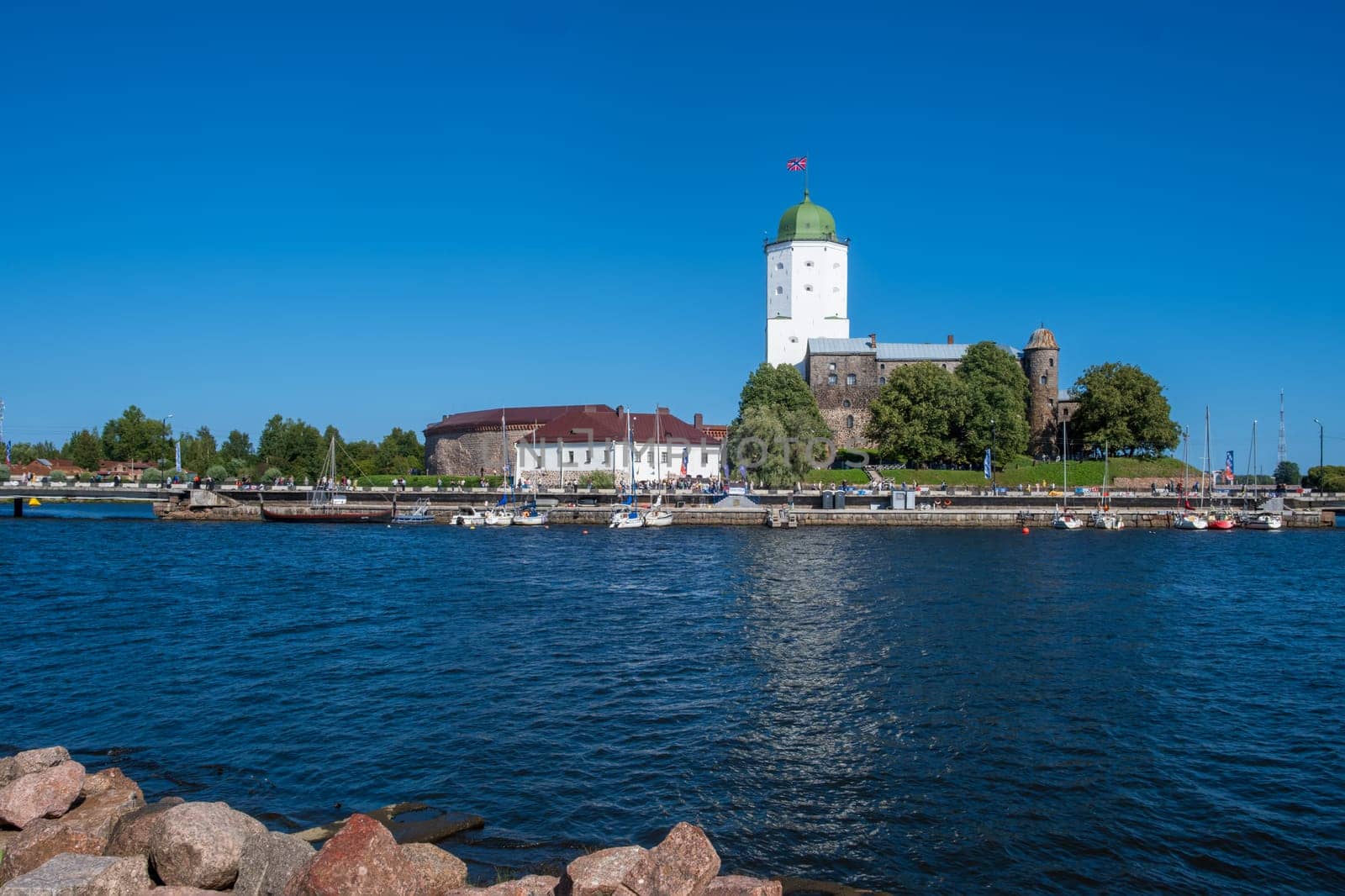 Vyborg, Leningrad region - 08.25.2024. Historic medieval knight's castle surrounded by calm blue waters. Boats are moored nearby and people are walking along the embankment.