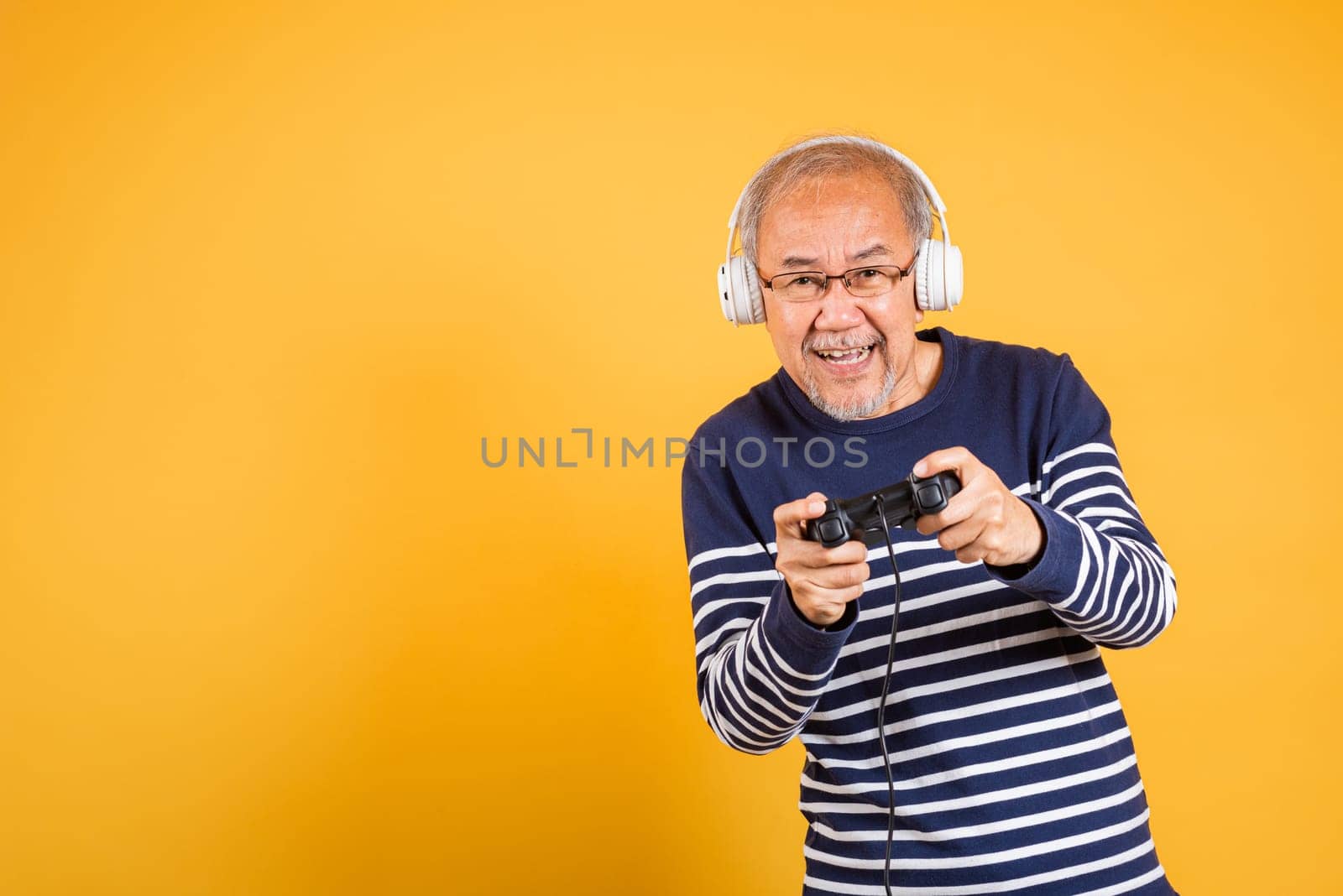 Portrait Asian smiling old man with headphones holding game controller studio shot isolated yellow background, senior man pensioner feeling winning playing a video game with joystick console