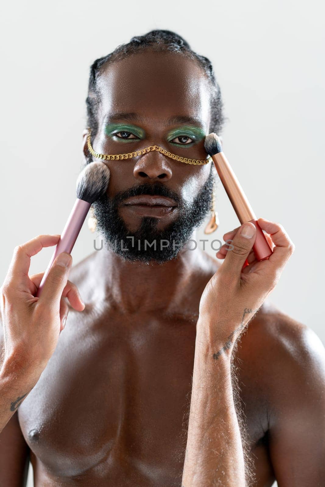 Portrait of African American gay man applying makeup with brushes held by a crop artist. Bearded man with bright eyeshadow and gold chain, looking at the camera