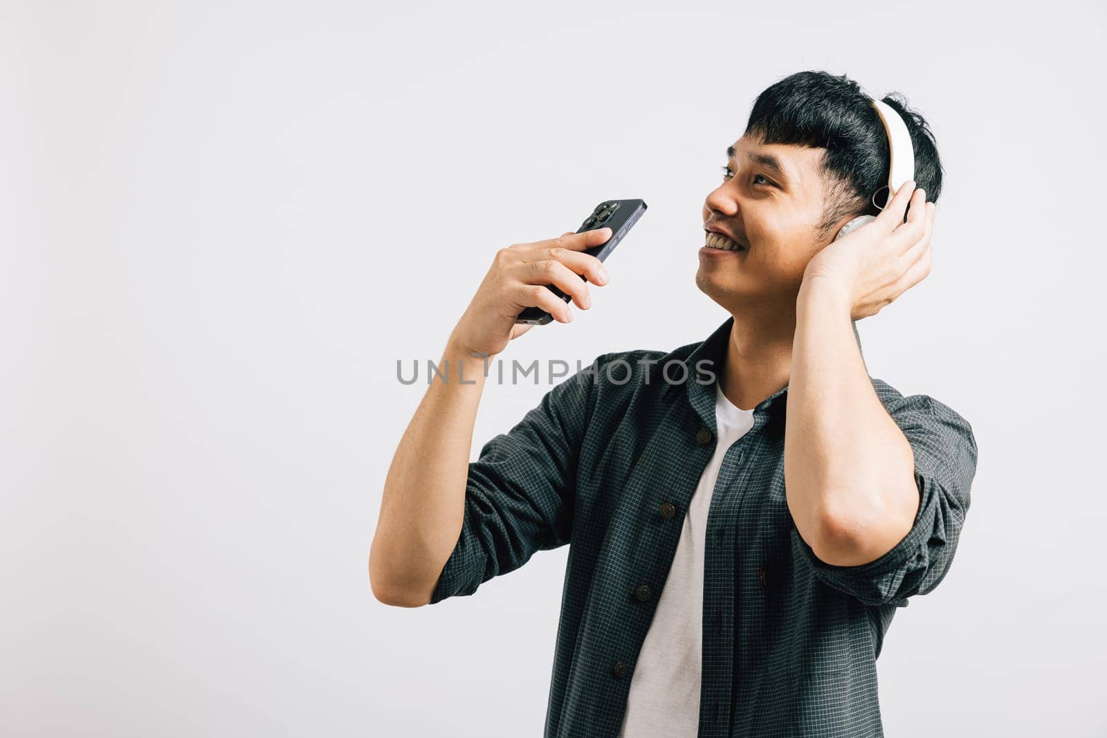 A young Asian man, positively immersed in karaoke, sings along to music on his smartphone with Bluetooth headphones. Studio shot isolated on white background, capturing his joy.