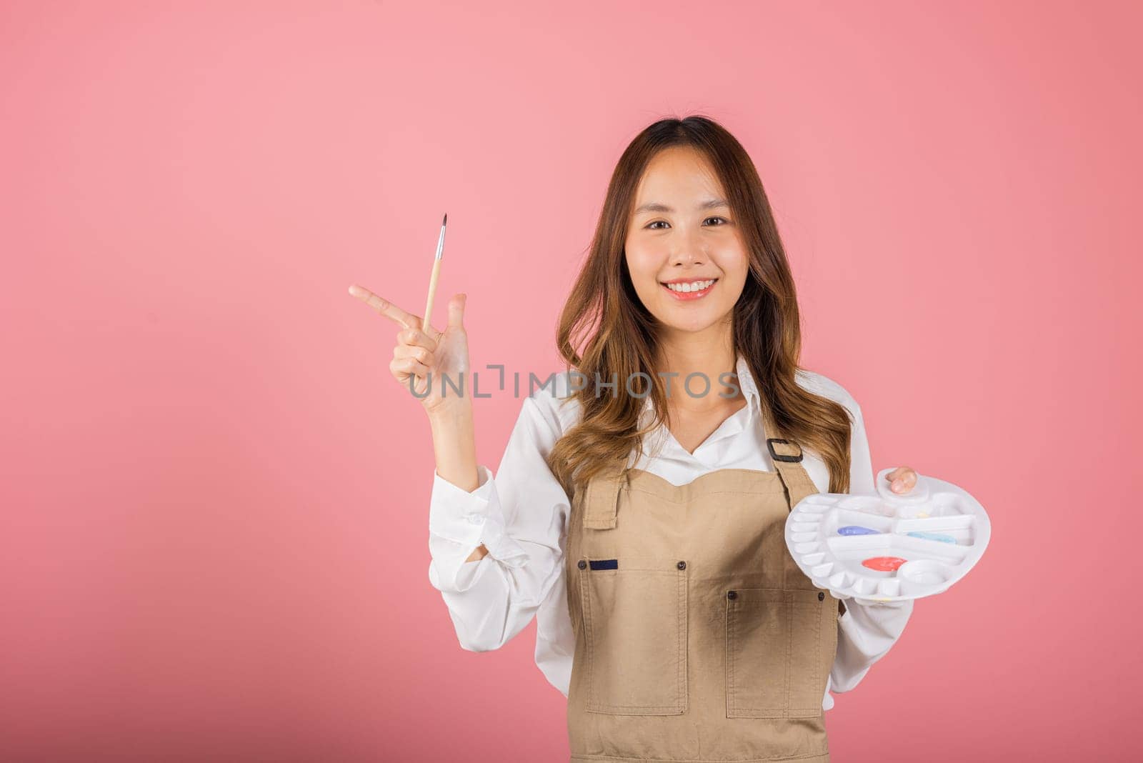 Asian beautiful young woman artist holding brush and paint palette, Happy female painting using paintbrush and palette with colors, studio shot isolated on pink background, Paintings and art equipment