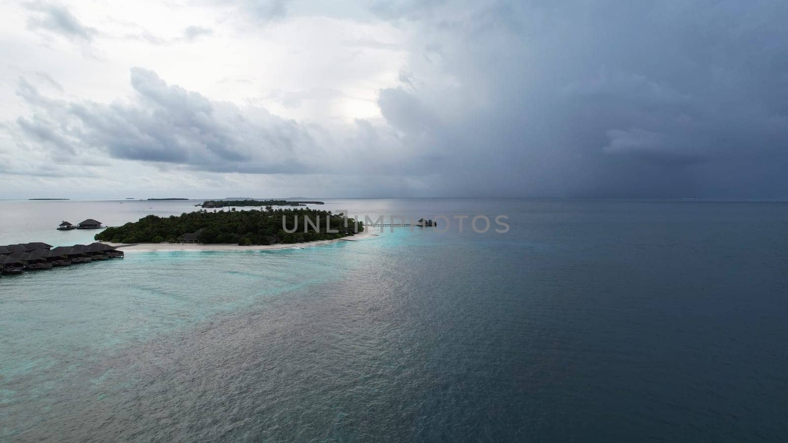 Aerial view of a luxury resort in the maldives with overwater bungalows and a pristine beach, as dark storm clouds gather in the distance, creating a dramatic contrast