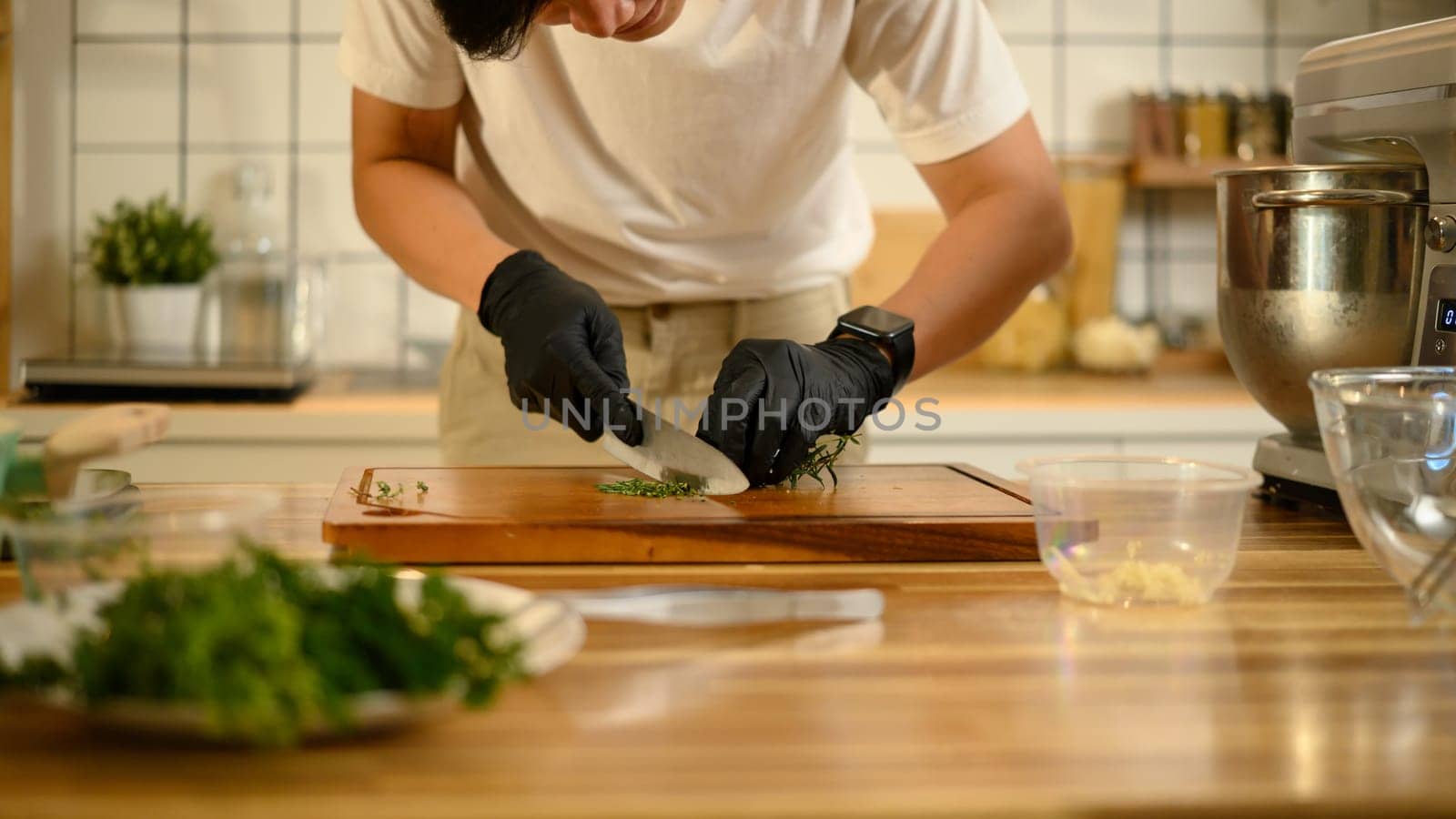 Man wearing black gloves chopping herbs on a wooden cutting board.