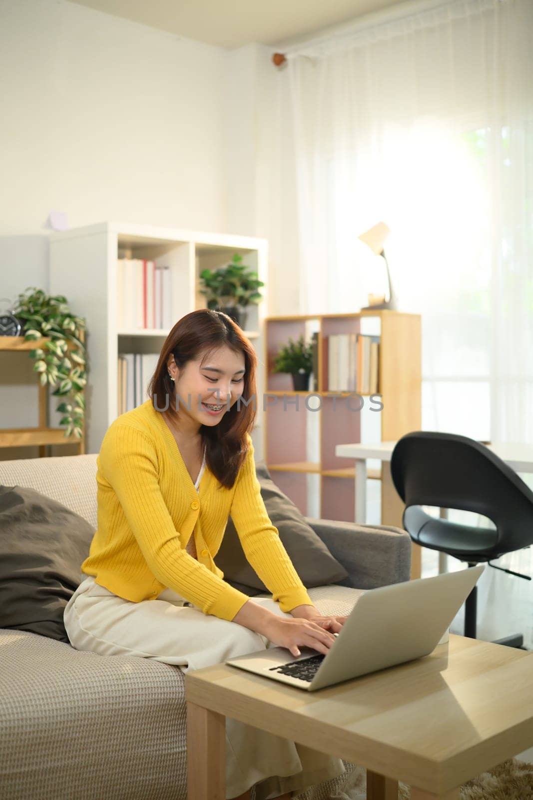 Young woman in yellow sweater working on her laptop in a bright living room.