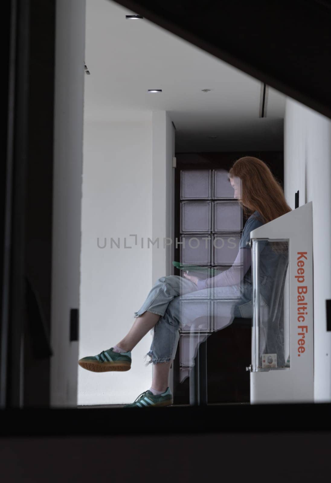 Newcastle, UK - Apr 12, 2025 - Looking through the glass at a long-haired young woman wearing T-shirt, jeans, sneakers sitting reading a book on bench in Baltic Flour Mills is Centre for Contemporary Art at Gateshead Quayside by the River Tyne. use it as your Wallpaper, Poster and Space for text, Selective focus.