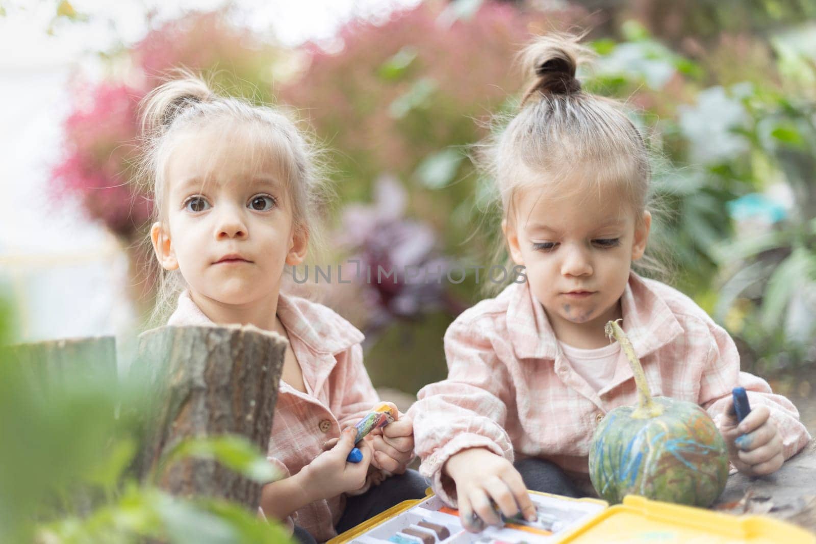 Two 3-year-old twin girls sit in a garden surrounded by bushes, drawing on a pumpkin with wax crayons. A creative and playful autumn moment highlighting childhood imagination