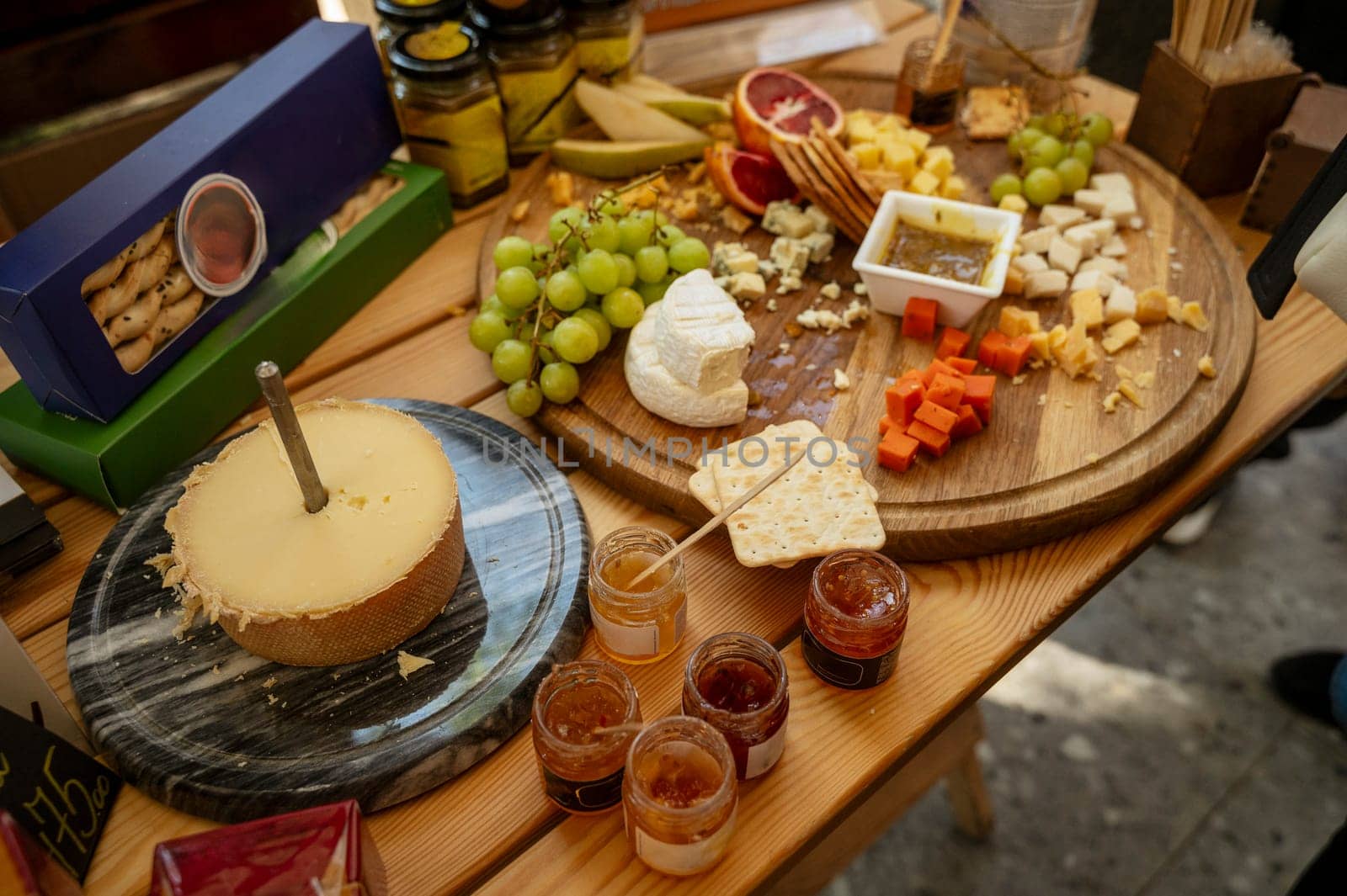Assorted cheeses on a round wooden board at a farmers market for tasting. Camembert cheese, a wheel of hard cheese, honey, grapes, crackers, bread, grapes, cheese slicers.