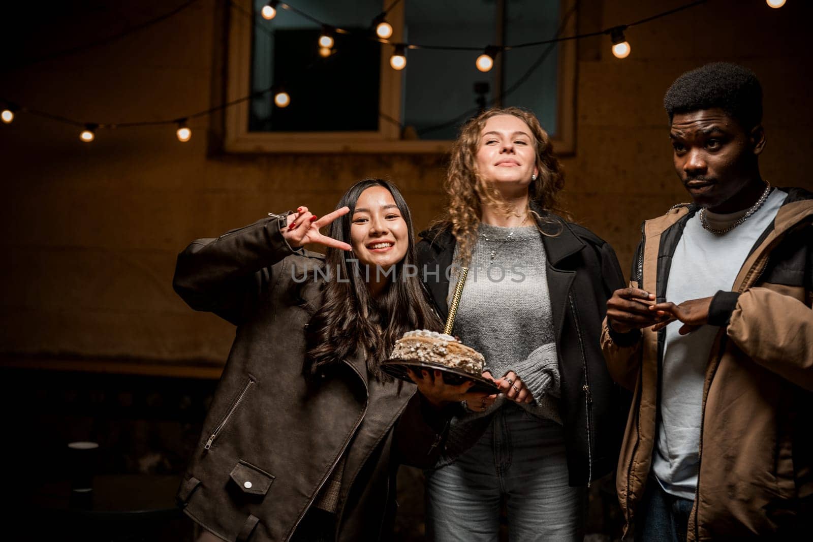Three friends enjoy a joyful evening together in a cozy space. One holds a delicious cake, while the others smile and pose for a fun moment, embodying the spirit of celebration and friendship.