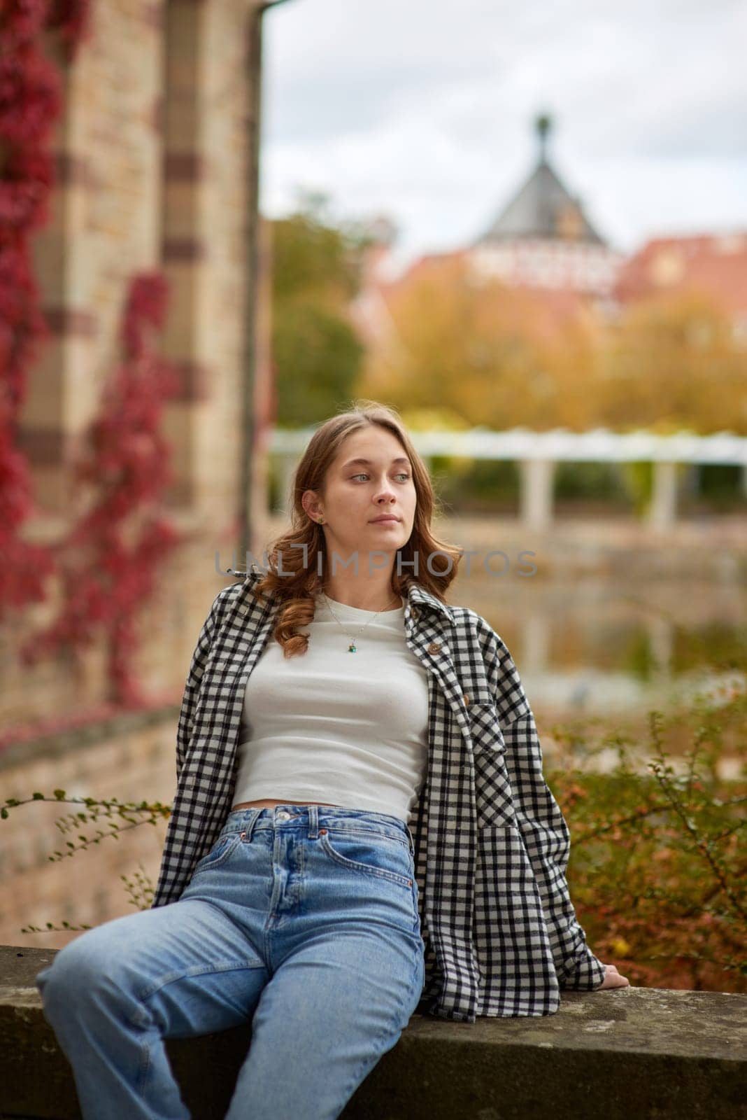 A stylish young woman with wavy brown hair and blue eyes sits on a stone ledge in a historic European city park, wearing a trendy black-and-white plaid shirt, a white crop top, and classic blue jeans. The soft natural lighting highlights the warm autumn atmosphere with golden leaves, red ivy, and medieval architecture in the background. Her relaxed pose and thoughtful expression create a cozy and nostalgic mood, perfect for lifestyle branding, travel promotions, autumn fashion editorials, and advertising seasonal collections. This image captures the essence of autumn in a historic European city, ideal for marketing campaigns, blog posts, and social media content related to travel, fashion, and seasonal themes. Stylish young woman in casual autumn outfit relaxing in historic European city park with medieval architecture, golden leaves, and red ivy, showcasing cozy autumn atmosphere, lifestyle, and travel concepts.