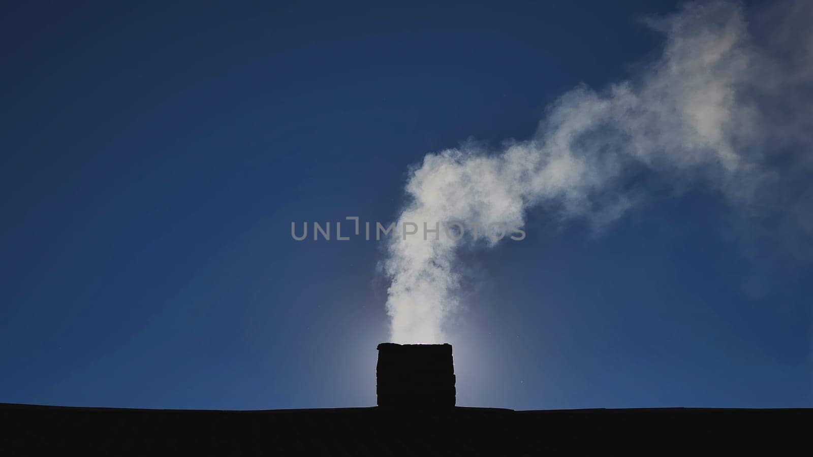 Billowing white smoke rising from chimney against clear blue sky, signaling winter heating or industrial emissions