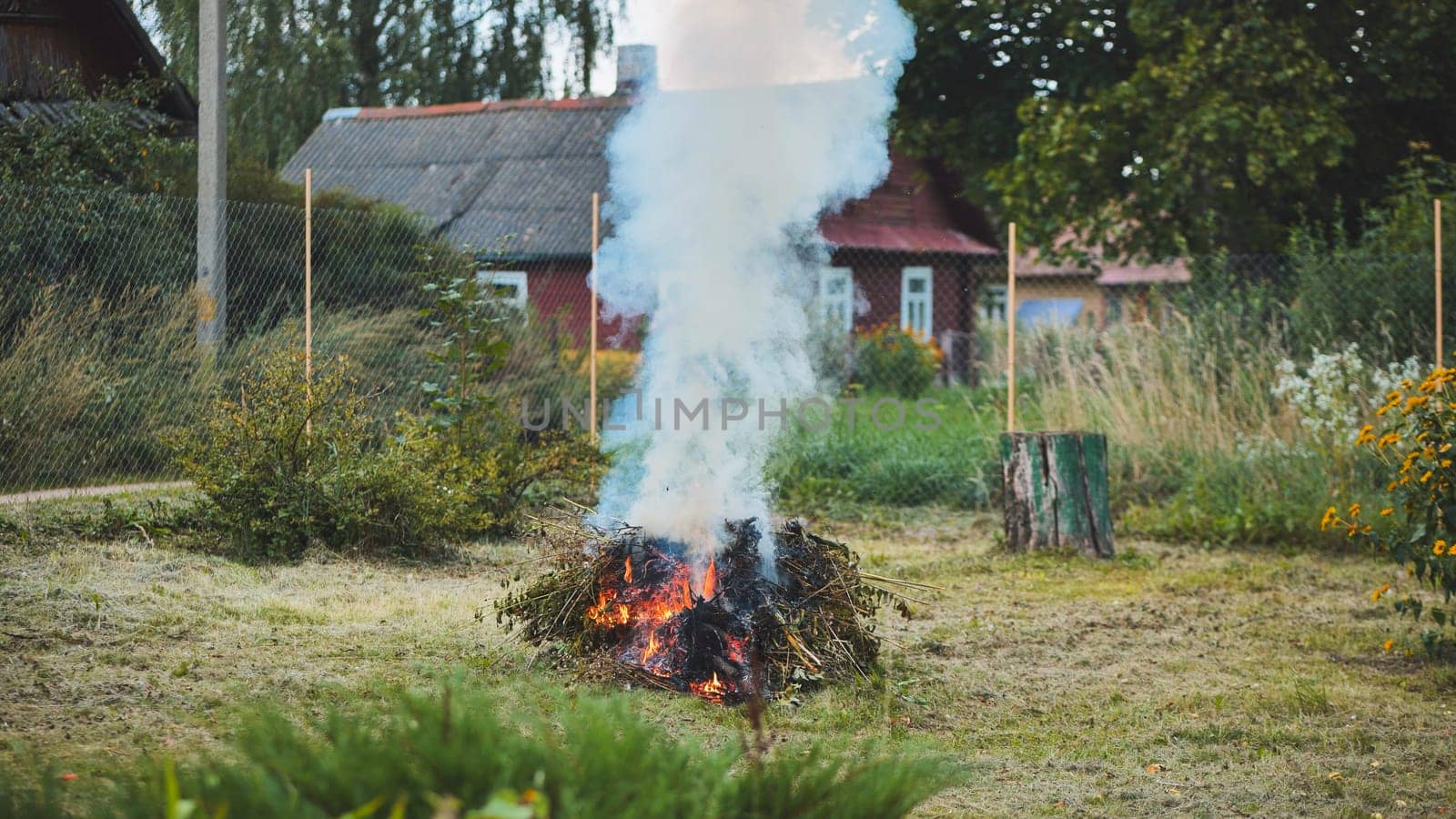 Pile of tree branches burning in a rural garden, producing a large plume of smoke, with a wooden house and fence in the background